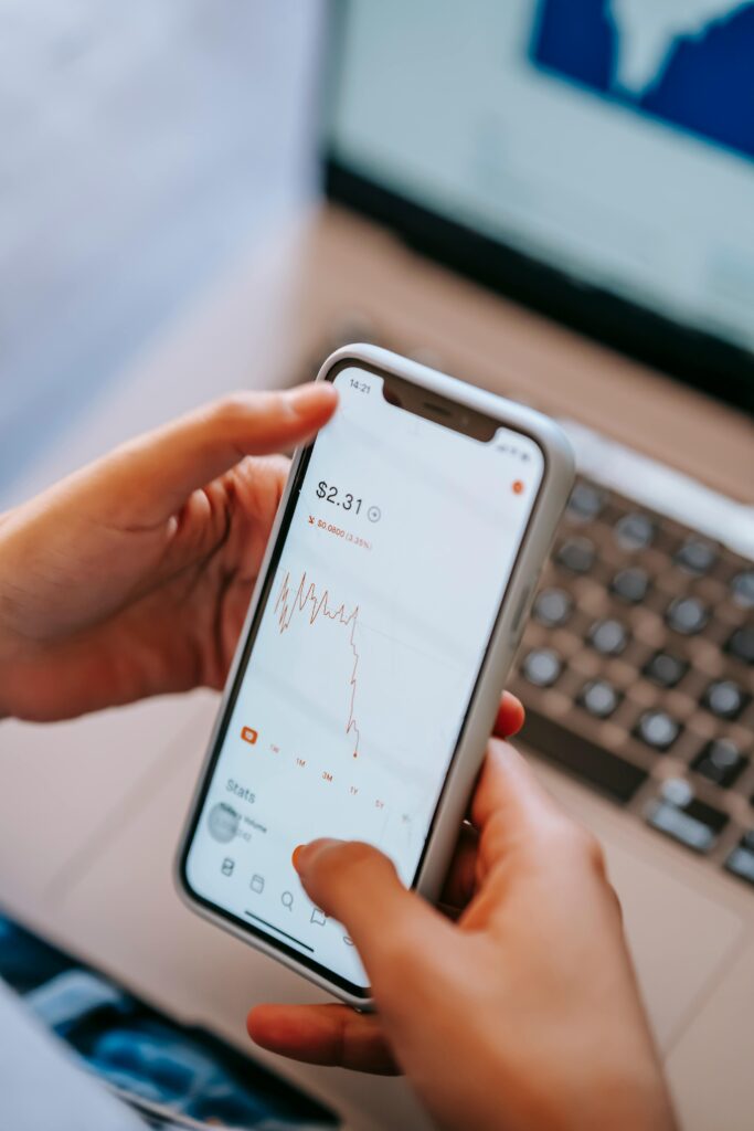 High angle of crop anonymous dealer using financial exchange application on cellphone with chart on screen against laptop